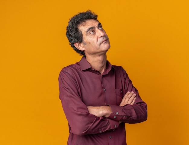 Senior man in purple shirt looking up with serious face puzzled with arms crossed standing over orange background