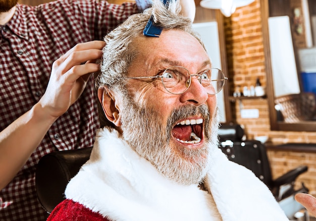 The senior man in Santa claus costume shaving his personal master at barber shop before Christmas