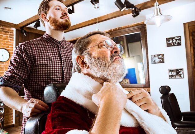 The senior man in Santa claus costume shaving his personal master at barber shop before Christmas