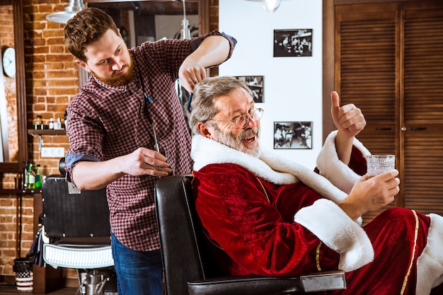 The senior man in Santa claus costume shaving his personal master at barber shop before Christmas