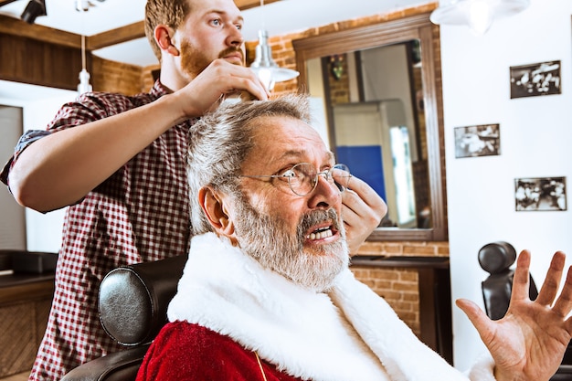 Foto gratuita l'uomo anziano in costume di babbo natale che rade il suo maestro personale al negozio di barbiere prima di natale