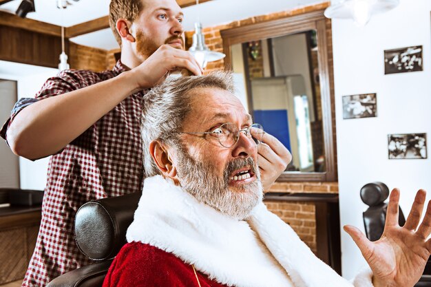 The senior man in Santa claus costume shaving his personal master at barber shop before Christmas