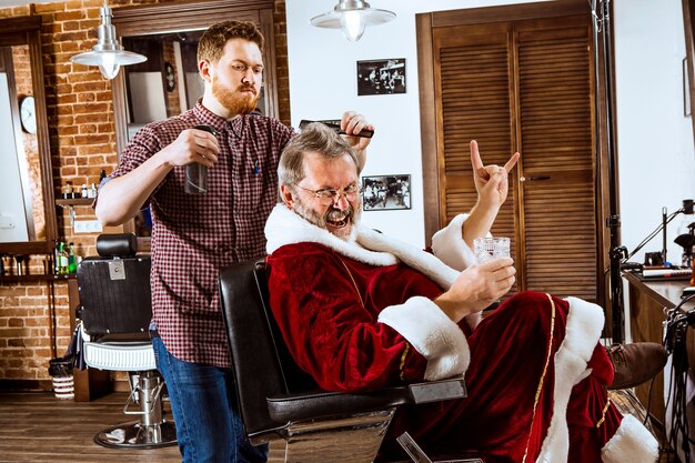 senior man in Santa claus costume shaving his personal master at barber shop before Christmas