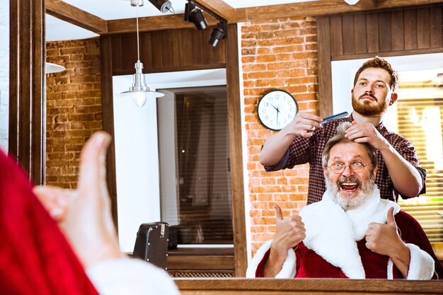 The senior man in Santa claus costume shaving his personal master at barber shop before Christmas