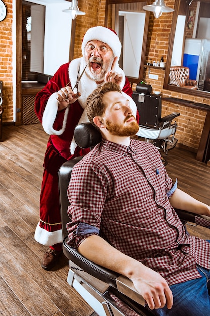 The senior man in santa claus costume working as personal master with scissors at barber shop before christmas