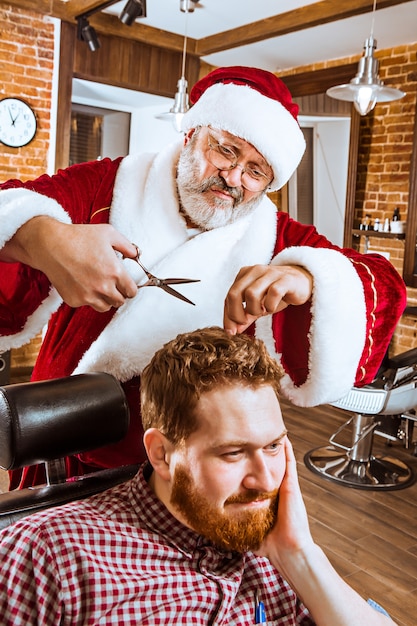 The senior man in Santa claus costume working as personal master with scissors at barber shop before Christmas