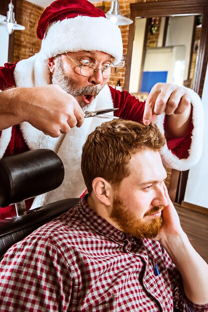 The senior man in Santa claus costume working as personal master with scissors at barber shop before Christmas