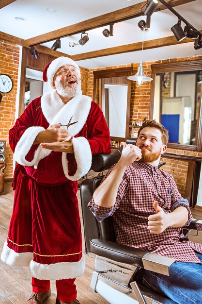 クリスマス前に理髪店でハサミで個人的なマスターとして働いているサンタクロースの衣装を着た年配の男性