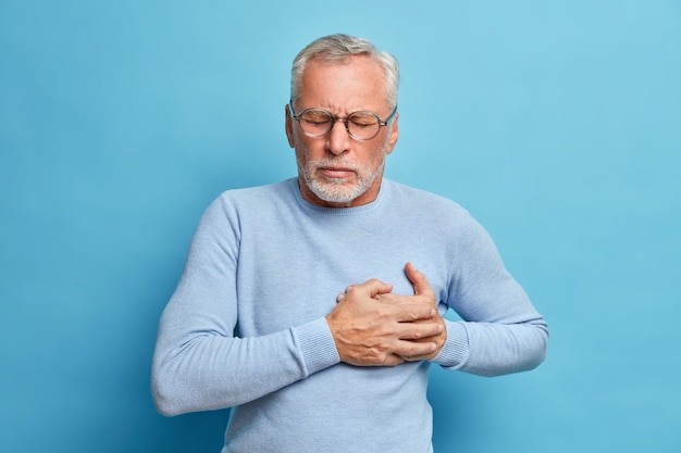 Senior man in spectacles presses hand to chest has heart attack suffers from unbearable pain closes eyes wears optical glasses poses against blue wall