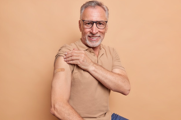 Senior man in spectacles shows plastered arm after getting coronavirus vaccine happy to feel safe and protected isolated over brown wall