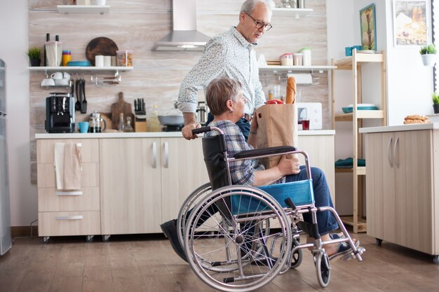Старший мужчина принимает продуктовый бумажный пакет от жены-инвалида в инвалидной коляске. Зрелые люди со свежими овощами с рынка. Проживание с инвалидом с нарушениями ходьбы