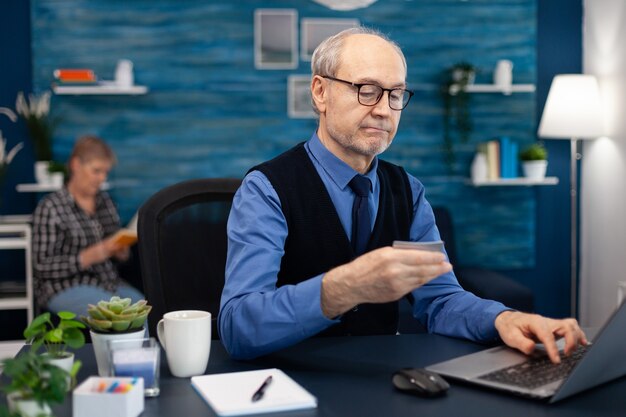 Senior man using credit card to check bank account. Elderly man checking online banking to make shppping payment looking at laptop while wife is reading a book sitting on sofa.