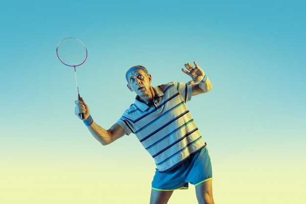 Senior man wearing sportwear playing badminton on gradient background, neon light. Caucasian male model in great shape stays active. Concept of sport, activity, movement, wellbeing, confidence.