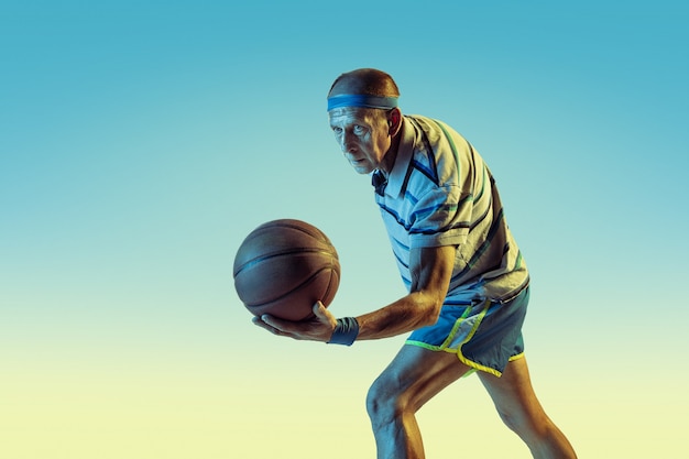 Uomo anziano che indossa abbigliamento sportivo che gioca a basket su sfondo sfumato, luce al neon. il modello maschile caucasico in ottima forma rimane attivo. concetto di sport, attività, movimento, benessere, fiducia.