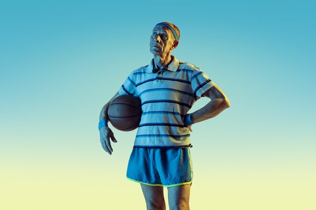 Senior man wearing sportwear playing basketball on gradient background, neon light.