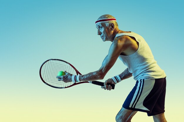 グラデーションの背景にネオンの光でテニスをするスポーツウェアを着た年配の男性。美しい体型の白人男性モデルは、アクティブでスポーティなままです。スポーツ、活動、動き、幸福、自信のコンセプト。