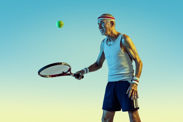 Senior man wearing sportwear playing tennis on gradient