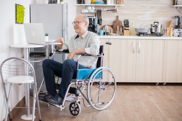 Старший мужчина в инвалидной коляске, используя ноутбук на кухне. Инвалид старший мужчина в инвалидной коляске, имея видеоконференцию на ноутбуке на кухне. Парализованный старик и его жена проводят онлайн-конференцию.