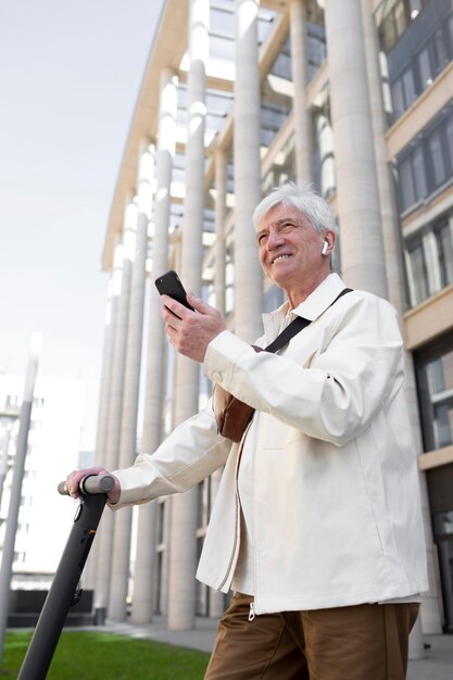 Старший мужчина с электросамокатом в городе с помощью смартфона и наушников