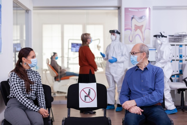 Senior man with face mask discussing with woman patient in stomatology clinic in waiting room, keeping social distancing during global pandemic with coronavirus