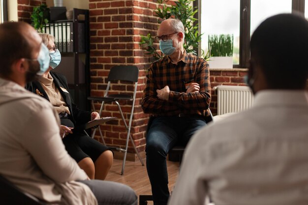 Senior man with face mask talking to people in circle at aa meeting, to cure alcohol addiction at support group therapy. Person having discussion with psychologist about problems.