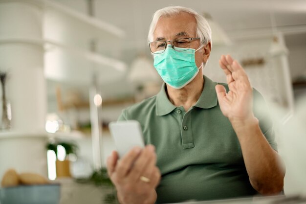 Senior man with face mask waving while making video call over smart phone at home