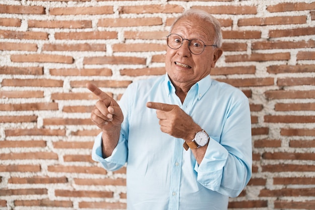 Senior man with grey hair standing over bricks wall pointing aside worried and nervous with both hands concerned and surprised expression