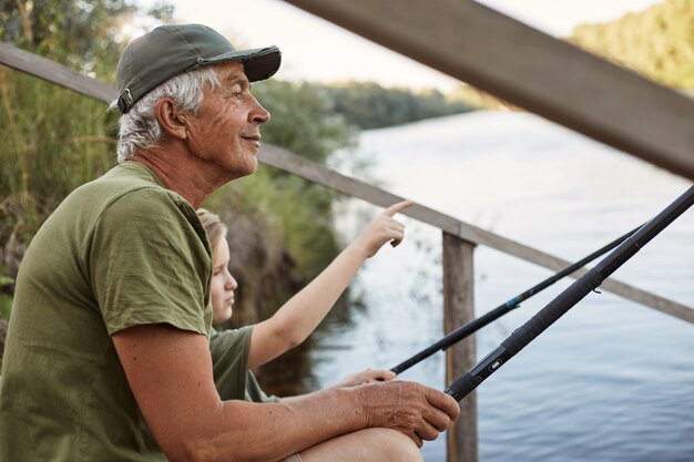 釣り竿を手に木製のポンツーンの上に座って、美しい自然、彼の指で何かを指している少年を楽しんでいる彼の孫を持つ年配の男性。