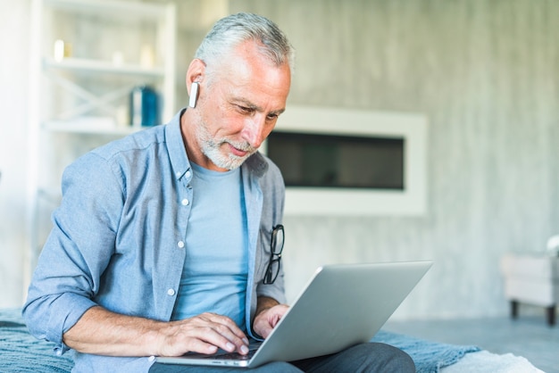 Senior man with wireless bluetooth sitting on bed using laptop