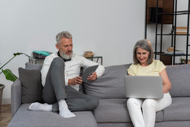 Senior man and woman at home on the couch using laptop and tablet