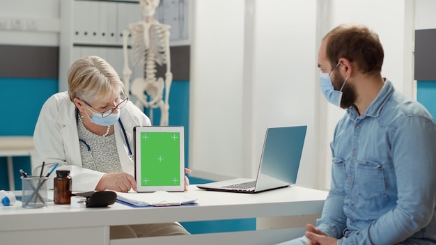 Старший медик и пациент используют зеленый экран на цифровом планшете во время пандемии ковида 19. Доктор показывает пациенту пустой макет фона с изолированным хромакеем copyspace на шаблоне. Съемка со штатива.