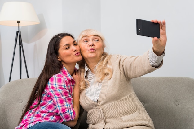 Senior mother and adult daughter sitting on sofa taking selfie on mobile phone