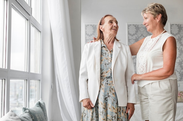 Free photo senior mother and daughter looking at each other while standing near window