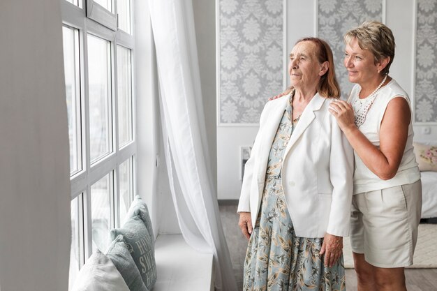 Senior mother and daughter looking through window at home