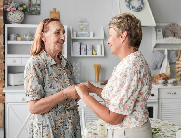 Senior mother and mature daughter holding hand together in kitchen