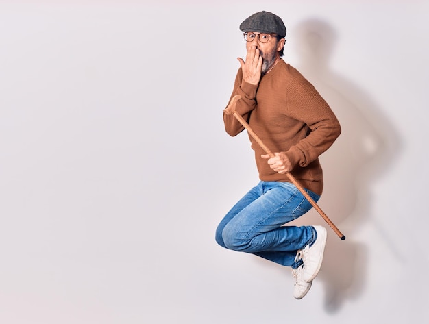 Senior old man wearing beret and glasses surprised with hand on mouth Jumping with excited expression holding walking stick over isolated white background