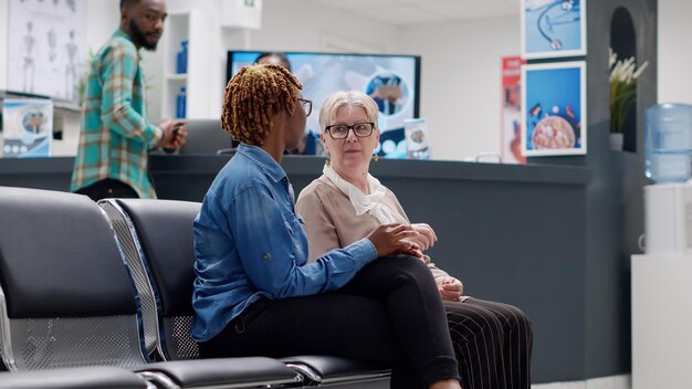 Senior patient and african american woman talking in waiting area at medical facility reception. Diverse people sitting in hospital waiting room to attend healthcare checkup visit. Handheld shot.