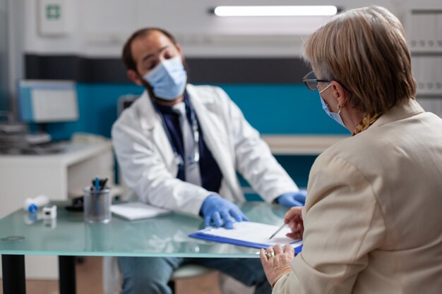 Senior patient signing checkup papers to receive prescription medicine at consultation with doctor. Man with face mask asking for signature to give health care treatment after examination.