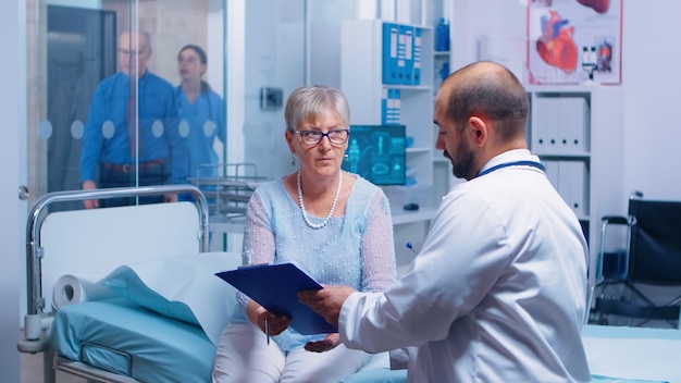 Senior patient signing medical decisions form sitting on hospital bed in modern private clinic. Doctor with clipboard, nurse working in backgorund. Healthcare medical medicinal system documents contra