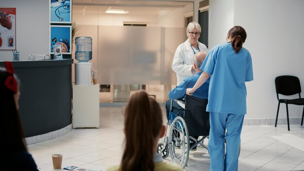 Senior physician talking to retired man with disability at hospital reception, preparing to do medical consultation. Wheelchair user in waiting room lobby attending checkup appointment.