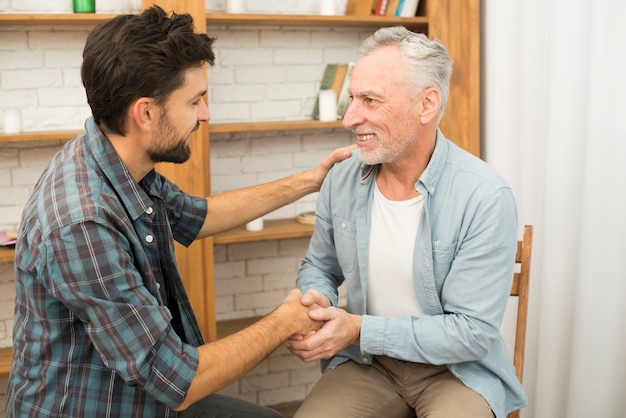 Senior positive man shaking hands with young happy guy in room