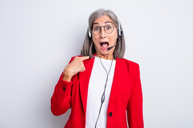 Senior pretty woman looking shocked and surprised with mouth wide open, pointing to self. telemarketer concept