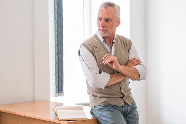 Senior professor holding glasses near desk in classroom