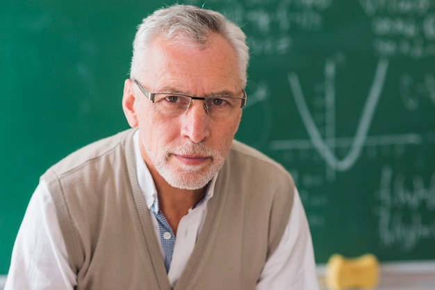 Foto gratuita professore senior che guarda l'obbiettivo contro la lavagna con esempio di matematica