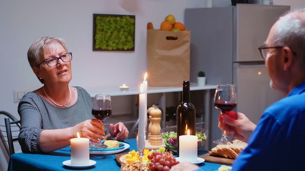Старшие расслабленные пары обедают и выпивают бокалы красного вина вместе на кухне дома. Пожилые люди пенсионного возраста наслаждаются трапезой, отмечают годовщину в столовой.