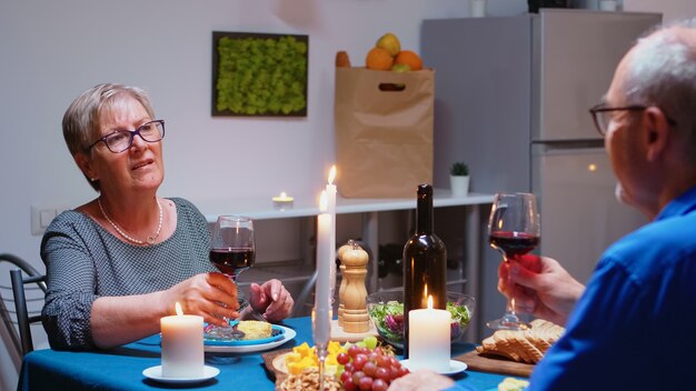 自宅のキッチンで一緒に夕食を食べ、赤ワインのグラスを飲むシニアリラックスしたカップル。食堂で結婚記念日を祝い、食事を楽しんでいるお年寄りの老人。