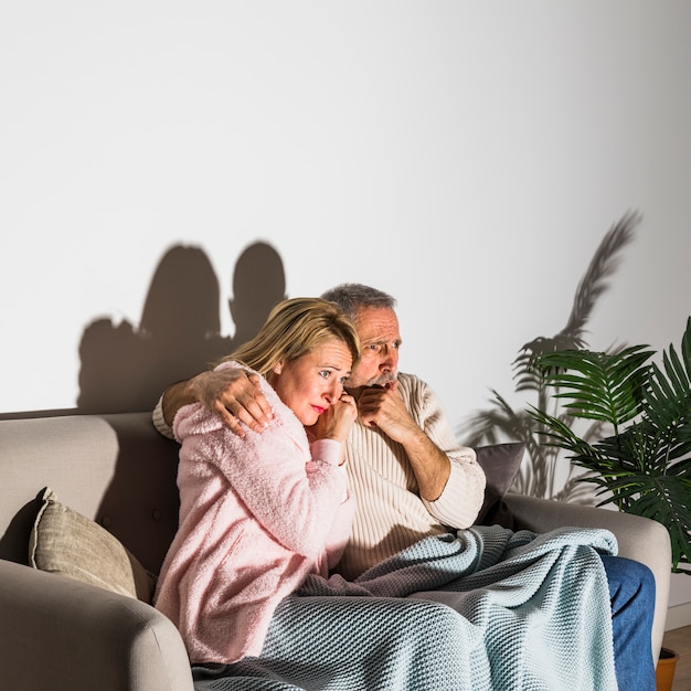 Senior scared man hugging with woman and watching TV on sofa