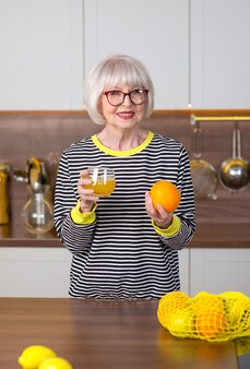 キッチン​に​立っている​間​レモネード​の​レモン​を​保持している​ストライプ​の​セーター​の​シニア​笑顔​の​女性