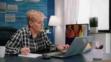 Free photo senior stylish woman taking notes in notebook while using laptop at home. old freelancer writing details on book while working from workspace in living room, checking financial project of company