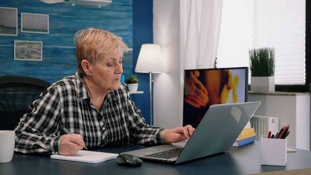 Senior stylish woman taking notes in notebook while using laptop at home. Old freelancer writing details on book while working from workspace in living room, checking financial project of company